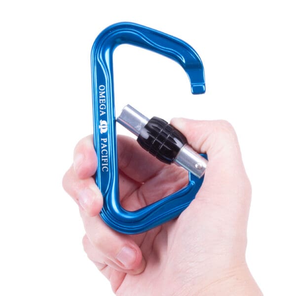 A hand holding a blue Custom Omega Pacific Standard D Aluminum Keylock Screw-Lok NFPA (5 Pack) with a black locking mechanism. The carabiner is being displayed against a plain white background.